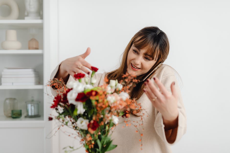 A smiling woman with light brown hair and bangs, wearing a beige sweater, is holding a vibrant bouquet of fresh flowers close to her face. The bouquet includes red, white, and orange flowers with gree