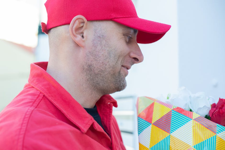 A man wearing a red cap and red jacket holds a colorful floral gift bag, filled with white and pink flowers, while smiling and looking at the flowers. The floral arrangement appears fresh with vibrant