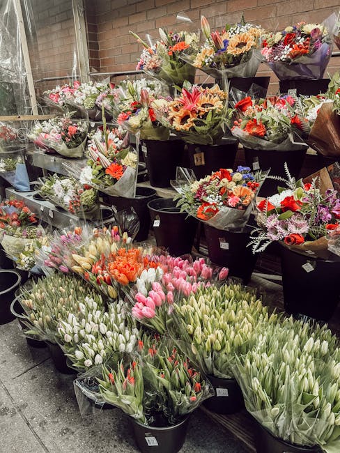 A display of numerous flower bouquets and bunches arranged in black buckets and wrapped in decorative paper, featuring a variety of fresh flowers including tulips in shades of pink, white, and red; ge