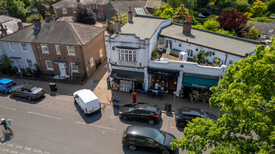 An aerial view of a small shopping street with several parked cars along the curb, including a white van and dark-colored vehicles. On the right side, a white, slightly curved building houses East She