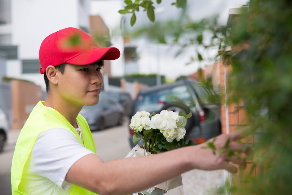 A young male florist wearing a red cap and a high-visibility yellow vest is arranging a bouquet of white flowers, likely hydrangeas, outdoors on a residential street. The bouquet is held in his left h